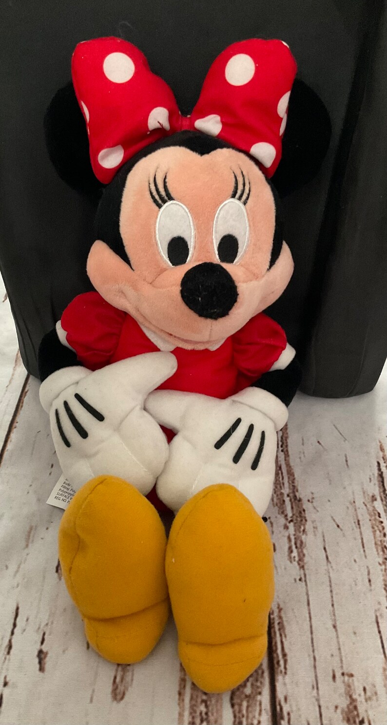 May include: A plush Minnie Mouse doll. The doll has a red dress, a red bow with white polka dots, black ears, and yellow shoes. The doll's hands are white with black gloves. The doll is sitting on a wooden surface.