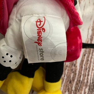 Puede incluir: Un juguete de peluche Mickey Mouse rojo y negro con una cara amarilla y guantes blancos. El juguete tiene una etiqueta blanca pegada que dice "Disney store" y "www.disneystore.com".