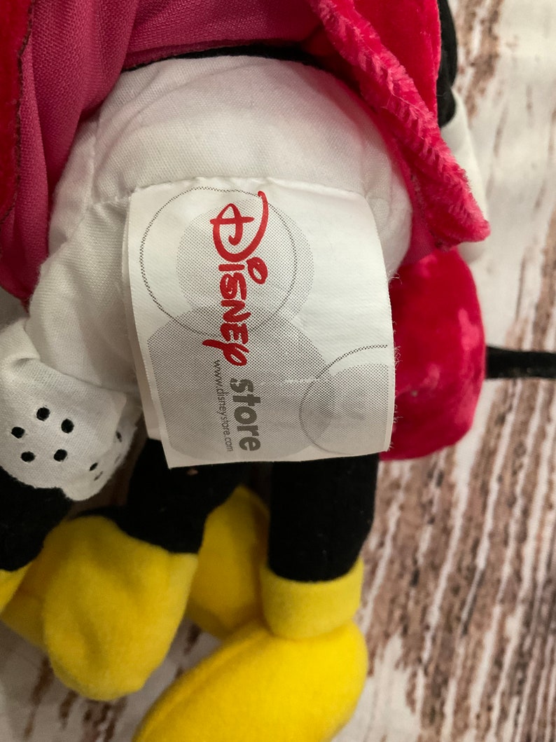 Puede incluir: Un juguete de peluche Mickey Mouse rojo y negro con una cara amarilla y guantes blancos. El juguete tiene una etiqueta blanca pegada que dice "Disney store" y "www.disneystore.com".