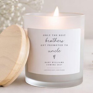 May include: A frosted glass candle with a lit wick and a light-colored wooden lid. The white label features the black text: ONLY THE BEST brothers GET PROMOTED TO uncle, a small heart icon, and the text BABY WILLIAMS COMING 2027.