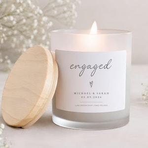 May include: A lit, frosted glass candle with a wooden lid, labeled "engaged" with a heart and names. The candle is surrounded by white flowers. The date 05.09.2026 is printed below the names. The candle is hand-poured.