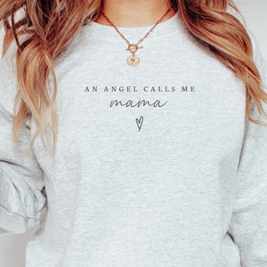 Può includere: Una felpa grigia con il testo "An angel calls me mama" e un piccolo cuore.