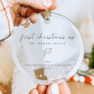 Könnte beinhalten: Runder, klarer Weihnachtsschmuck mit goldenem Band. Der Schmuck trägt den Text "first christmas as DR. DANIEL DAVIS" und eine Grafik einer Doktorhut-Kappe. Darunter steht "PHD BRIGHTON HILLS COLLEGE 2025".