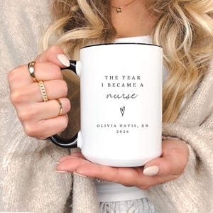 May include: White ceramic mug with a black handle and rim. The mug features the text "THE YEAR I BECAME A nurse" with a heart symbol and the name "OLIVIA DAVIS, RN 2026". The mug holds approximately 444 ml.