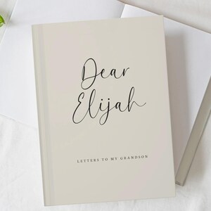 May include: A light beige hardback book with the text "Dear Elijah" in a black cursive script and "LETTERS TO MY GRANDSON" in a smaller, simple black font. The book is open, revealing blank white pages, and is positioned on a white fabric surface.