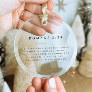 Könnte beinhalten: Ein kreisförmiges Ornament aus klarem Acryl mit dem schwarz aufgedruckten Text ROMANS 8:28 und dem Vers And we know that for those who love God all things work together for good, for those who are called according to his purpose, mit einem goldenen Band.