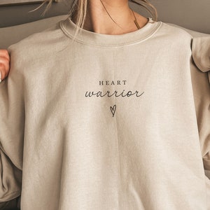 May include: A beige sweatshirt with the text "HEART warrior" and a small heart symbol printed on the front.
