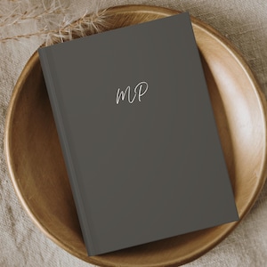 May include: A dark gray journal with the initials "MP" written in white on the front cover. The journal is resting on a wooden tray.