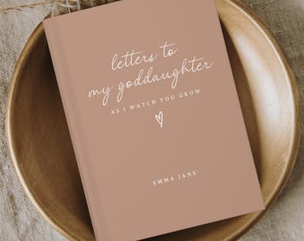 Op de afbeelding: Een hardcover boek met de titel "Letters to my Goddaughter" in witte letters op een stoffige roze kaft. Het boek ligt in een houten kom, met de naam "Emma Jane" onderaan.