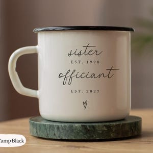 May include: A 10oz white enamel camping mug with a black rim and handle. The front features black text reading sister EST. 1998, officiant EST. 2027, and a small heart symbol. The mug is positioned on a round green stone coaster.