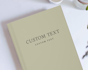 Puede incluir: Un libro de tapa dura color verde salvia con el texto impreso en negro CUSTOM TEXT seguido de una línea más pequeña de CUSTOM TEXT. El libro está colocado sobre una superficie lisa junto a una pequeña taza de cerámica blanca y un florero.