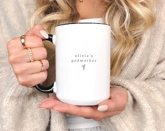 Può includere: Tazza in ceramica bianca con bordo e manico neri. La tazza ha il testo "olivia's godmother" con un piccolo simbolo di cuore.
