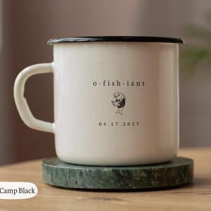 Puede incluir: Una taza de camping de esmalte blanco con borde y asa negros. El frente presenta el texto o-fish-iant sobre un pequeño gráfico negro de un pez con esmoquin sosteniendo un libro, con la fecha 04.17.2027 impresa en negro debajo del pez.