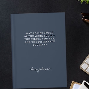 Könnte beinhalten: Ein marineblaues Notizbuch mit dem Text "MAY YOU BE PROUD OF THE WORK YOU DO, THE PERSON YOU ARE, AND THE DIFFERENCE YOU MAKE" und dem Namen "chris johnson" in Weiß. Es liegt auf einer schwarzen Oberfläche mit Tastatur, Brille und Pflanze.