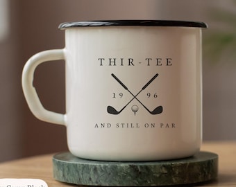 May include: A white enamel camp mug with a black rim and handle. The front features black text reading THIR-TEE 1996 AND STILL ON PAR, with a graphic of two crossed golf clubs and a golf ball. The mug is labeled as a 10oz Enam Camp Black.