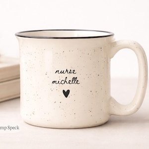 May include: Cream ceramic mug with black speckles and a black rim. The mug features the text "nurse michelle" and a black heart. The mug has a capacity of approximately 385 ml.