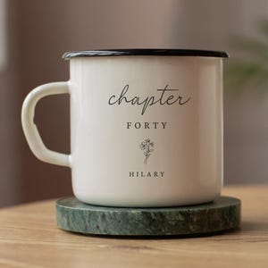 May include: White enamel mug with a black rim and handle, resting on a green marble coaster. The mug features the words "chapter FORTY" and the name "HILARY" in black script, along with a small floral illustration.