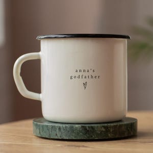 Puede incluir: Una taza de esmalte blanco con borde y asa negros, sobre un posavasos de mármol verde. La taza presenta el texto "anna's godfather" con un símbolo de corazón debajo. La taza está sobre una superficie de madera.
