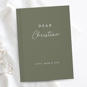 May include: A sage green hardback book with white text on the front cover. The text reads DEAR Christian in a mix of serif and script fonts, followed by the text LOVE, MOM & DAD at the bottom. The book is positioned on a white fabric surface.