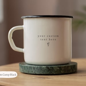 Op de afbeelding: Een witte emaille kampeermok met een zwarte rand en handvat. De mok heeft de tekst "your custom text here" en een hartsymbool. De mok staat op een groene marmeren onderzetter. De mok is 284 ml.