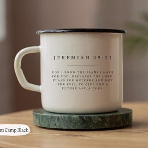 Może przedstawiać: Biały emaliowany kubek kempingowy z czarną obwódką i uchwytem, ​​na zielonym spodku. Kubek zawiera tekst "JEREMIASZ 29:11" i werset biblijny. Kubek mieści około 296 ml.
