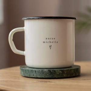 Puede incluir: Una taza de esmalte blanco con borde y asa negros, sobre un posavasos de mármol verde. La taza tiene el texto "nurse michelle" con un símbolo de corazón. La taza está sobre una superficie de madera.