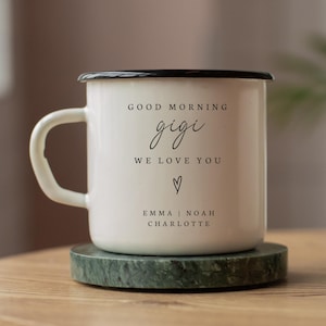 Puede incluir: Taza de esmalte blanco con borde y asa negros, sobre un posavasos de m&aacute;rmol verde. La taza presenta el texto "GOOD MORNING gigi WE LOVE YOU" con un coraz&oacute;n y los nombres Emma, Noah y Charlotte.