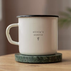 Może przedstawiać: Biały kubek emaliowany z czarną obwódką i uchwytem. Kubek ma napis "olivia's auntie" z małym symbolem serca nadrukowanym na nim. Kubek stoi na zielonej marmurowej podkładce.