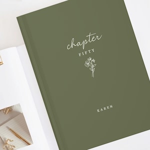 Puede incluir: Un diario verde oliva con las palabras "chapter FIFTY" en escritura blanca y una ilustración floral. El nombre "KAREN" está impreso en la parte inferior. El diario está abierto, revelando páginas en blanco y un clip dorado.
