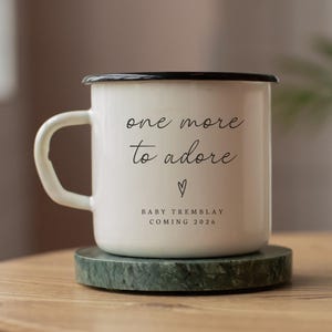 Può includere: Una tazza smaltata bianca con bordo e manico neri, su un sottobicchiere di marmo verde. La tazza presenta la scritta "one more to adore", un cuore e "BABY TREMBLAY COMING 2026".