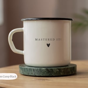 Peut inclure: Mug de camping en émail crème avec un bord et une anse noirs. Le mug porte l'inscription "MASTERED IT!" et un symbole de cœur noir. Le mug est posé sur un dessous de verre en marbre vert. Contenance : environ 296 ml.