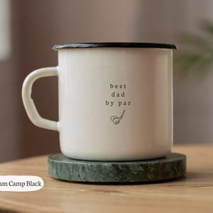 May include: A white enamel camp mug with a black rim and handle. The mug features the text "best dad by par" with a golf club and ball graphic. The mug is 10 oz and sits on a green marble coaster.