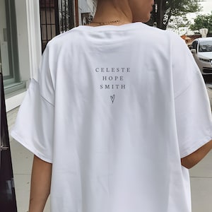 May include: White t-shirt with the text "CELESTE HOPE SMITH" and a heart symbol printed in black. The shirt has short sleeves and a relaxed fit. The person is wearing a gold necklace.