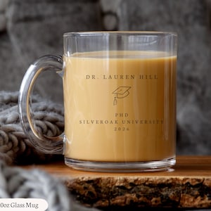 Könnte beinhalten: Ein klarer Glasbecher, gefüllt mit einer hellbraunen Flüssigkeit. Der Becher hat einen gebogenen Griff und zeigt den Text "DR. LAUREN HILL", eine Grafik einer Graduierungskappe, "PHD SILVEROAK UNIVERSITY 2026". Der Becher ist mit "10oz Glass Mug" beschriftet.