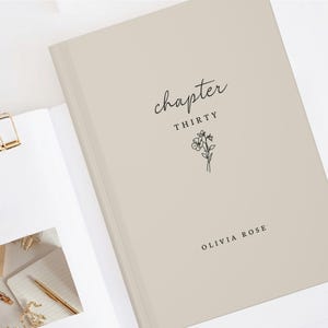 May include: A beige hardcover journal with the words "chapter thirty" and a floral illustration. The name "Olivia Rose" is printed at the bottom. A gold binder clip and other stationery items are visible.