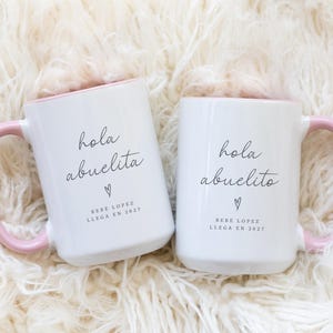 May include: Two white ceramic mugs with pink handles and rims. The mugs have the text "hola abuelita" and "hola abuelito" in black script, with a heart symbol and the text "BEBE LOPEZ LLEGA EN 2027".