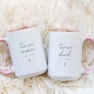 May include: Two white ceramic mugs with pink handles and interior rims. One mug reads "twin mom" and the other reads "twin dad" in black script. Both mugs have "EST. 2026" and a heart symbol below the text.