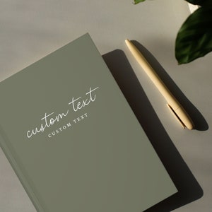 Peut inclure: Un carnet vert sauge avec le texte "custom text" et "custom text" en lettres blanches. Un stylo beige clair est posé sur une surface blanche à côté du carnet.