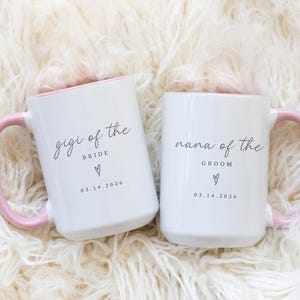 May include: Two white ceramic mugs with pink handles and rims. One mug reads "gigi of the bride" with a heart and the date 03.14.2026. The other mug reads "nana of the groom" with a heart and the date 03.14.2026. The mugs are on a fluffy white surface.