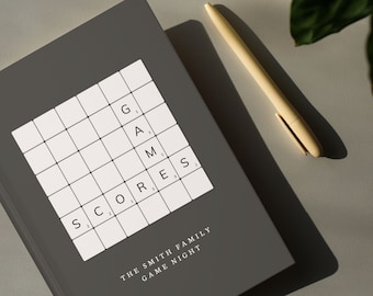 Puede incluir: Un cuaderno gris con una cuadrícula blanca y las palabras "SCORE", "G", "A", "M", "E", "S", "THE SMITH FAMILY GAME NIGHT" impresas en él. Un bolígrafo beige está sobre una superficie gris junto al cuaderno.