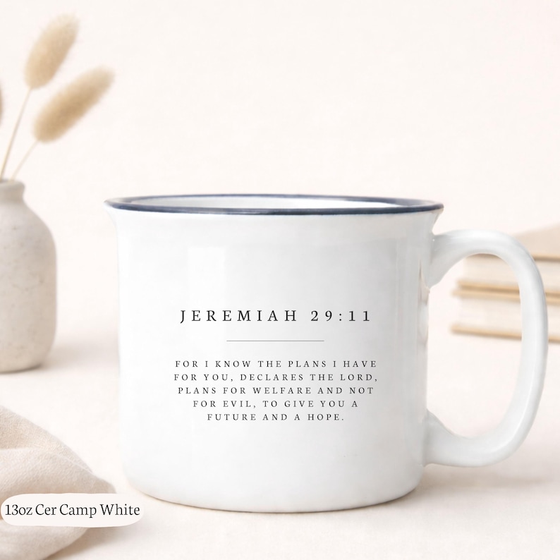 以下が含まれることがあります： 濃い青色の縁と取っ手が付いた白いセラミックマグカップ。「JEREMIAH 29:11」のテキストと長い引用句が特徴です。マグカップには「13oz Cer Camp White」と記載されています。