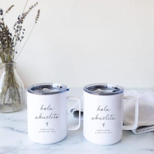 May include: Two white travel mugs with black text that reads "hola abuelito" and "hola abuelita" with a heart symbol. The text "BEBE LOPEZ LLEGA EN 2027" is also printed on the mugs. Each mug has a handle and a clear lid.