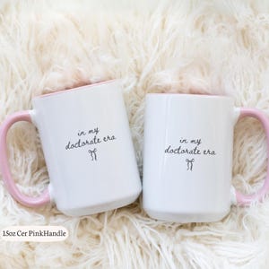 May include: Two white ceramic mugs with pink handles and interior. The mugs have the text "in my doctorate era" in black script. The mugs are sitting on a fluffy, white surface.