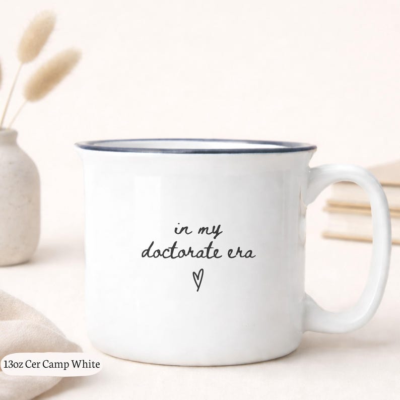 May include: White ceramic mug with a dark blue rim and handle. The mug features the text "in my doctorate era" with a heart symbol. The mug is 13oz and is labeled "Cer Camp White".
