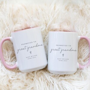 May include: Two white ceramic mugs with pink rims and handles. The mugs have the text "Promoted to Great Grandma" with a heart and the text "Baby Wilson June 2025".