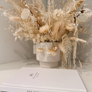 May include: A white book with the text "OUR JOURNEY TO YOU" and a rainbow graphic. A dried floral arrangement in a white vase is in the background. The arrangement includes beige and cream-colored flowers and grasses.