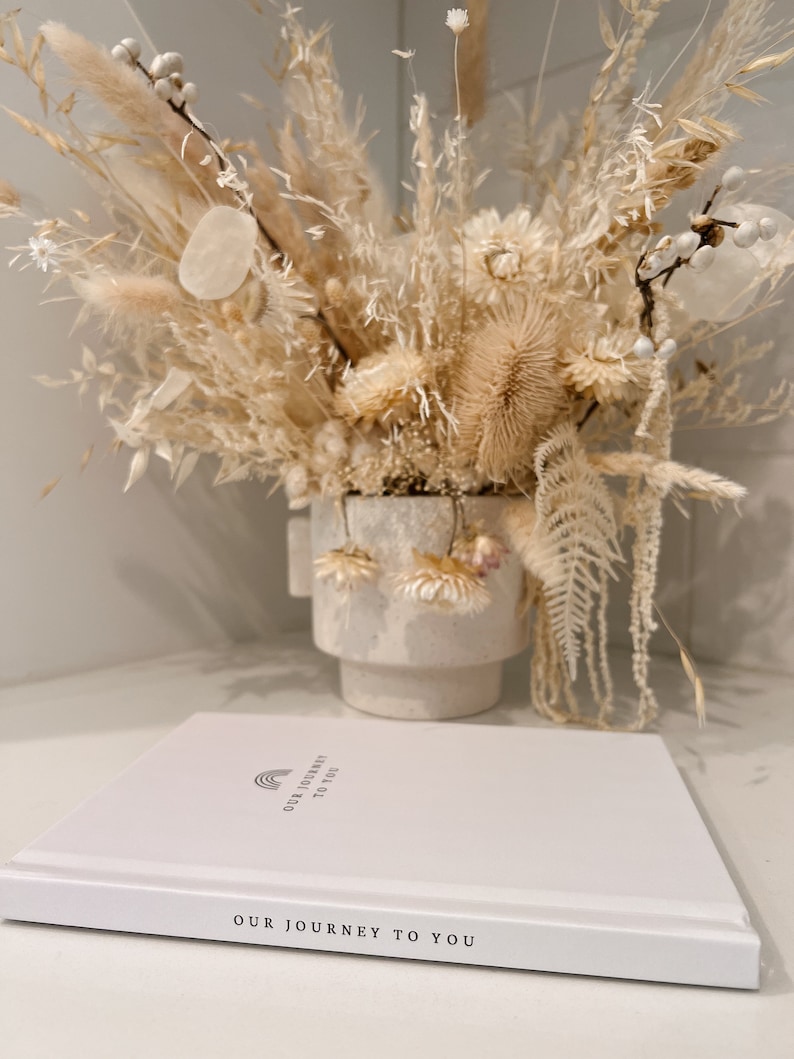May include: A white book with the text "OUR JOURNEY TO YOU" and a rainbow graphic. A dried floral arrangement in a white vase is in the background. The arrangement includes beige and cream-colored flowers and grasses.