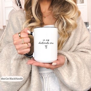 May include: White ceramic mug with a black handle and rim. The mug features the text "in my doctorate era" in a black script font, along with a small bow graphic. The mug is 15oz.