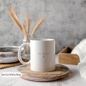 May include: A white ceramic mug with a handle, featuring the text "Dr. Miller" and a heart symbol. The mug is placed on a wooden coaster. The image also includes a wooden tray and decorative wheat stalks.