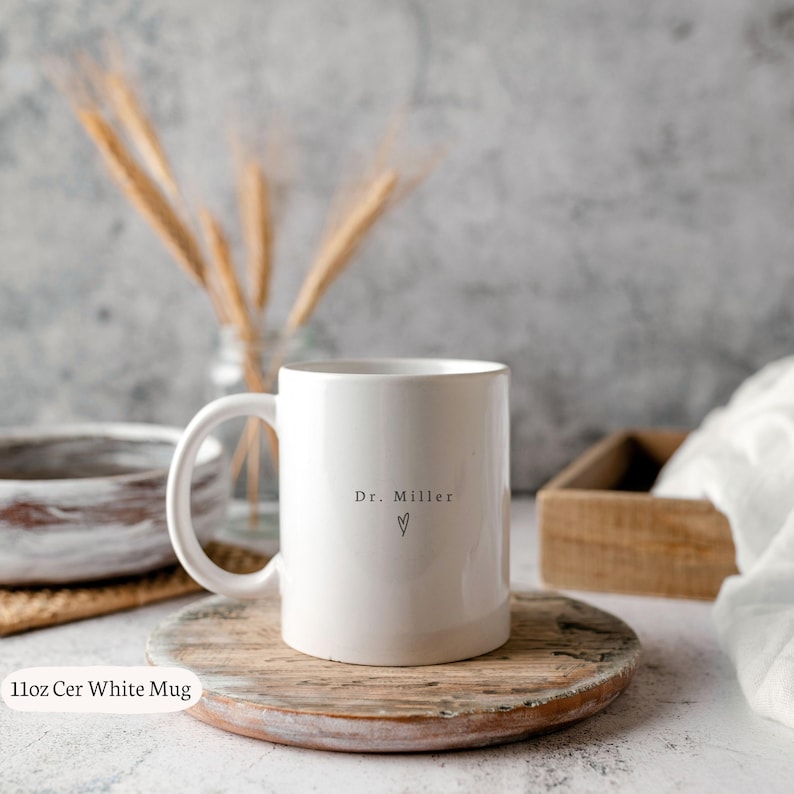 May include: A white ceramic mug with a handle, featuring the text "Dr. Miller" and a heart symbol. The mug is placed on a wooden coaster. The image also includes a wooden tray and decorative wheat stalks.